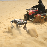 A DEEP Robotics Lynx M20 Pro wheel-legged quadruped robot traverses sandy desert terrain, kicking up dust, with a person riding a red ATV in the background also stirring up sand. The robot demonstrates its multi-terrain adaptability and autonomous navigation capabilities in extreme environments, equipped with features like LiDAR and an IP66 rating for industrial inspection and scientific exploration.