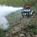 A DEEP Robotics Lynx M20 Pro wheel-legged quadruped robot, an industrial-grade and IP66-rated platform, is shown in a rugged, overgrown outdoor environment, actively spraying a white mist from a red tank mounted on its back. This demonstrates its capability for emergency response, such as firefighting or suppression, and autonomous operation in challenging terrains using features like LiDAR and advanced navigation.